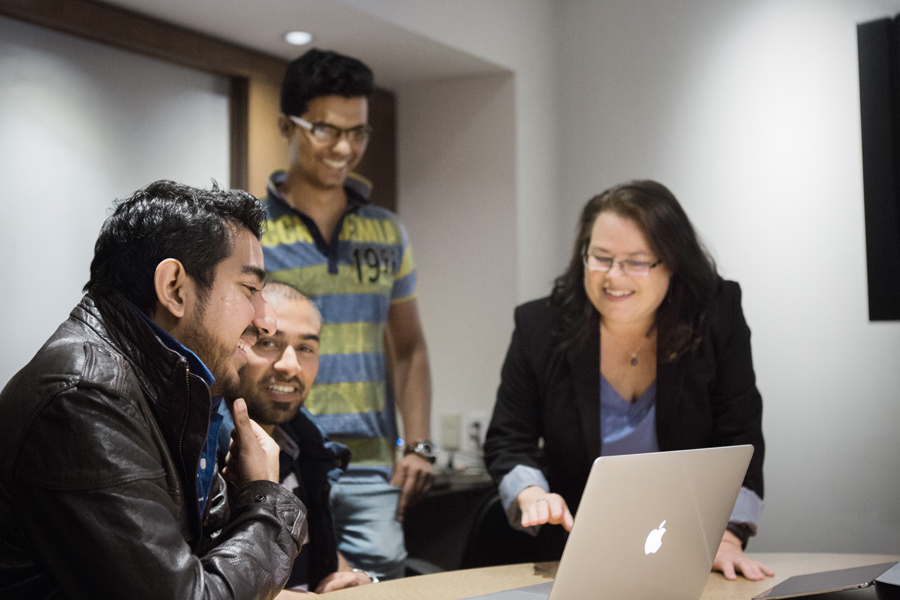 Smiling female staff member along with three male students with computer on desk.