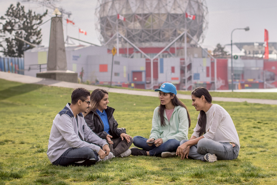 Against the backdrop of Science World, four New York Tech Vancouver students engage in a conversation while seated on a patch of grass.